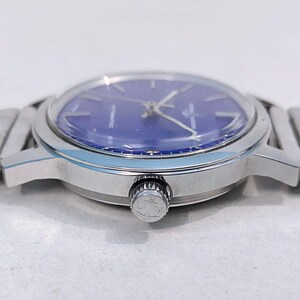 May include: A silver-toned wristwatch with a blue face. The watch has a round dial with silver hands and markers. The watch band is made of silver links. The watch is shown from a side angle.
