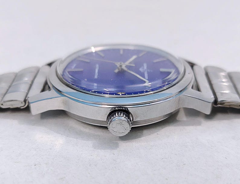 May include: A silver-toned wristwatch with a blue face. The watch has a round dial with silver hands and markers. The watch band is made of silver links. The watch is shown from a side angle.