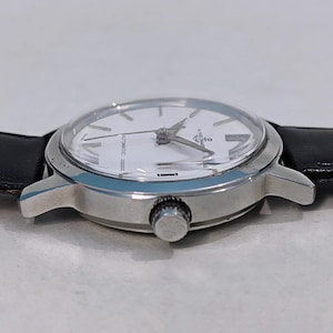 May include: A silver-toned wristwatch with a white face and black leather band. The watch has a round face with silver hands and markers. The watch is approximately 1.5 inches in diameter.