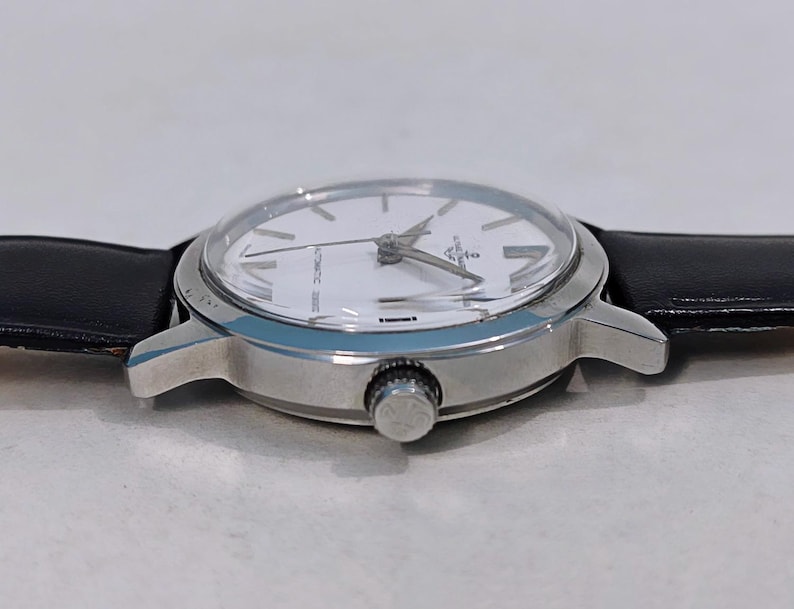 May include: A silver-toned wristwatch with a white face and black leather band. The watch has a round face with silver hands and markers. The watch is approximately 1.5 inches in diameter.