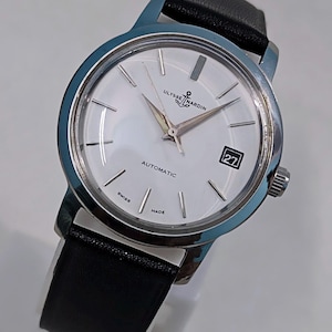 May include: A silver-toned Ulysse Nardin automatic wristwatch with a white face and black leather band. The watch face features silver hands and markers, a date window, and the words "Ulysse Nardin" and "Automatic". The watch is marked "Swiss Made".