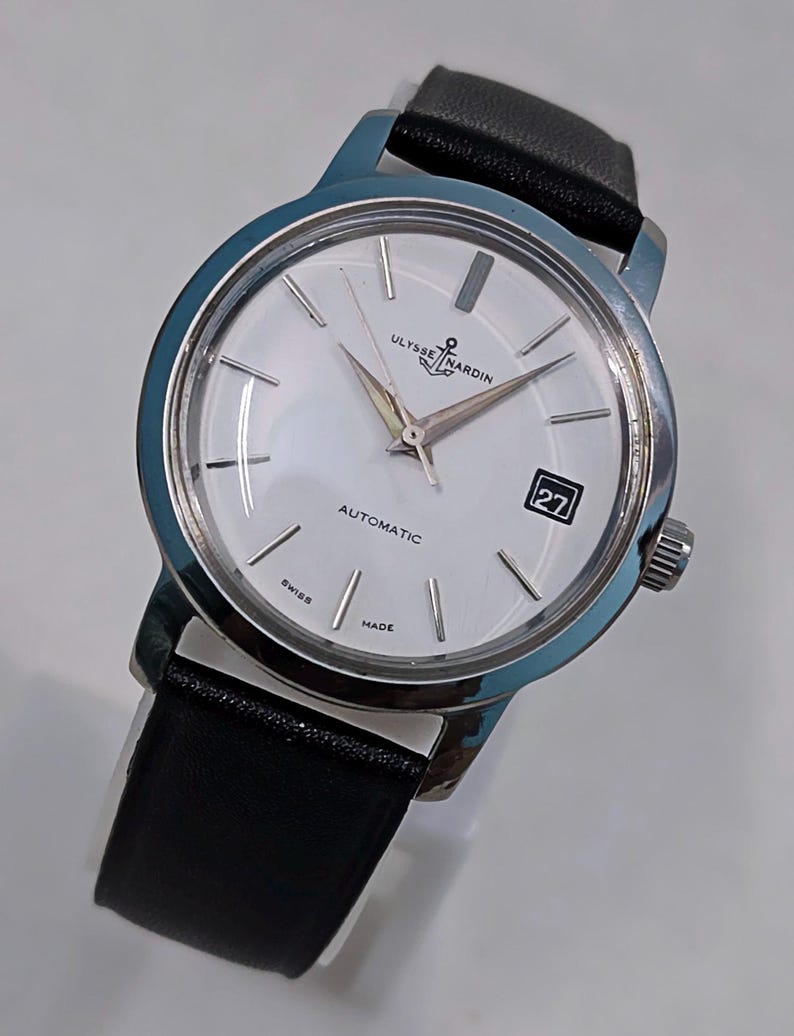 May include: A silver-toned Ulysse Nardin automatic wristwatch with a white face and black leather band. The watch face features silver hands and markers, a date window, and the words "Ulysse Nardin" and "Automatic". The watch is marked "Swiss Made".