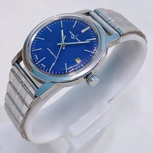 May include: A silver-toned Ulysse Nardin automatic watch with a deep blue face. The watch features a date window and a silver band. The watch is displayed on a white stand.