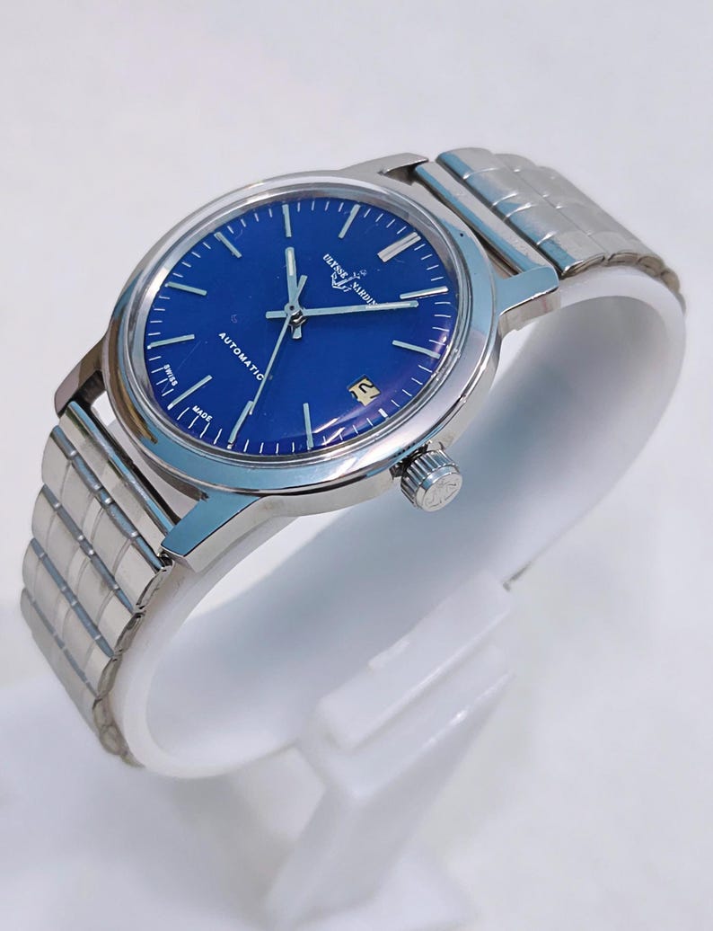 May include: A silver-toned Ulysse Nardin automatic watch with a deep blue face. The watch features a date window and a silver band. The watch is displayed on a white stand.