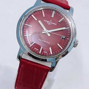 May include: A stainless steel Ulysse Nardin automatic watch with a burgundy dial and red leather strap. The watch face features silver hands and markers, with a date display at the 3 o'clock position. The watch is marked "Swiss Made".