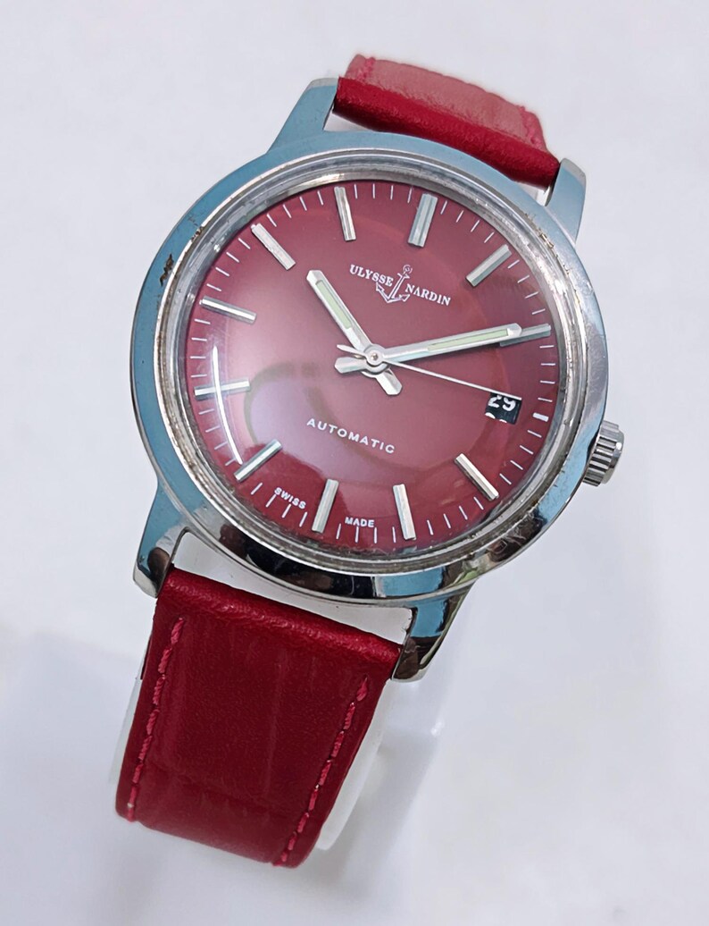 May include: A stainless steel Ulysse Nardin automatic watch with a burgundy dial and red leather strap. The watch face features silver hands and markers, with a date display at the 3 o'clock position. The watch is marked "Swiss Made".