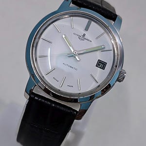 K&ouml;nnte beinhalten: Eine silberfarbene Ulysse Nardin Automatik-Armbanduhr mit wei&szlig;em Zifferblatt, schwarzem Lederarmband und Datumsanzeige. Die Uhr hat eine silberne L&uuml;nette und Zeiger mit leuchtend gr&uuml;nen Akzenten. Das Zifferblatt zeigt "AUTOMATIC", "SWISS" und "MADE".