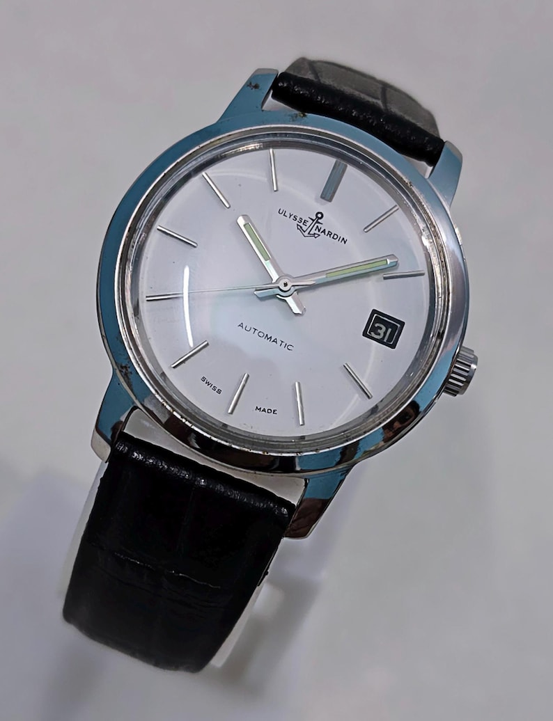 K&ouml;nnte beinhalten: Eine silberfarbene Ulysse Nardin Automatik-Armbanduhr mit wei&szlig;em Zifferblatt, schwarzem Lederarmband und Datumsanzeige. Die Uhr hat eine silberne L&uuml;nette und Zeiger mit leuchtend gr&uuml;nen Akzenten. Das Zifferblatt zeigt "AUTOMATIC", "SWISS" und "MADE".
