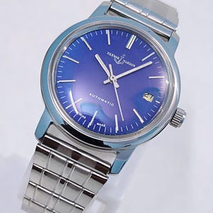 May include: A stainless steel Ulysse Nardin automatic watch with a blue dial. The watch features silver hands and markers, a date window at the 3 o'clock position, and a stainless steel bracelet. The watch face has the text "Ulysse Nardin", "Automatic", "Swiss Made".