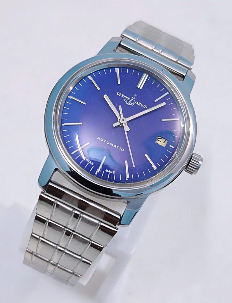 May include: A stainless steel Ulysse Nardin automatic watch with a blue dial. The watch features silver hands and markers, a date window at the 3 o'clock position, and a stainless steel bracelet. The watch face has the text "Ulysse Nardin", "Automatic", "Swiss Made".
