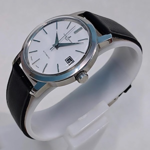 May include: A silver-toned wristwatch with a white face, black hands, and a date window at the 3 o'clock position. The watch has a black leather band and the text "ULYSSE NARDIN" is visible on the face. The watch is displayed on a white stand.