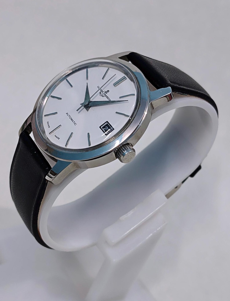May include: A silver-toned wristwatch with a white face, black hands, and a date window at the 3 o'clock position. The watch has a black leather band and the text "ULYSSE NARDIN" is visible on the face. The watch is displayed on a white stand.