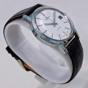 K&ouml;nnte beinhalten: Eine silberfarbene Armbanduhr mit wei&szlig;em Zifferblatt und schwarzem Lederarmband. Das Zifferblatt hat silberne Stundenmarkierungen, ein Datumsfenster und den Text "ULYSSE NARDIN AUTOMATIC SWISS MADE". Die Uhr wird auf einem wei&szlig;en St&auml;nder pr&auml;sentiert.