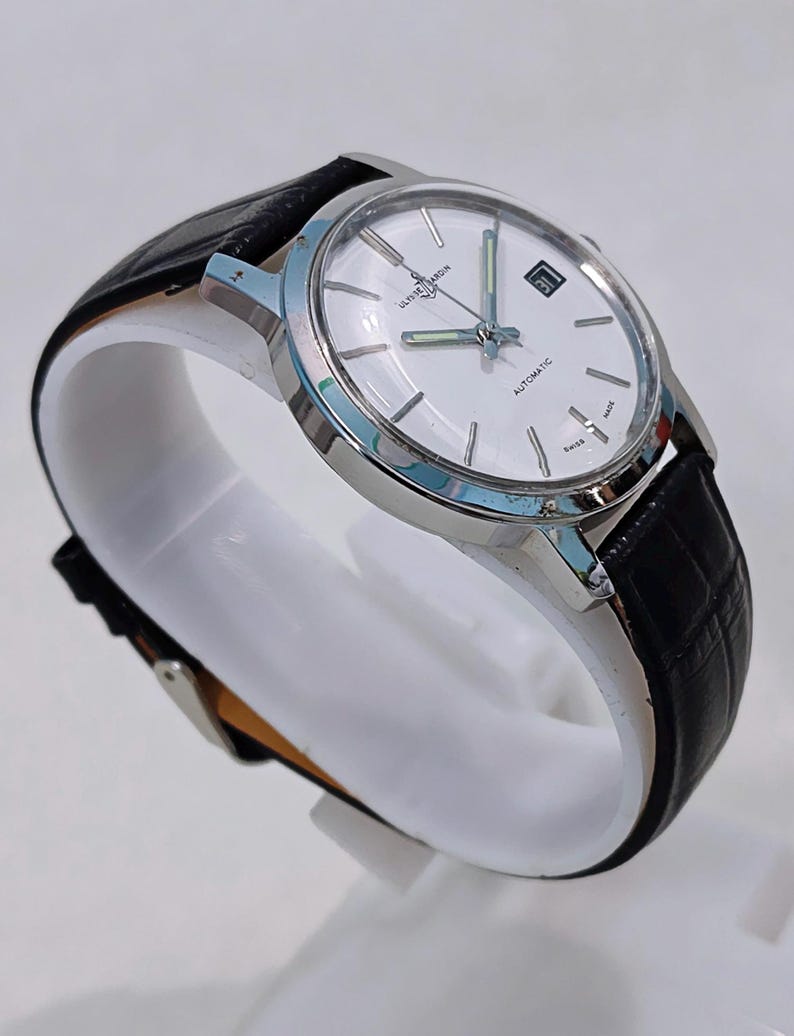 K&ouml;nnte beinhalten: Eine silberfarbene Armbanduhr mit wei&szlig;em Zifferblatt und schwarzem Lederarmband. Das Zifferblatt hat silberne Stundenmarkierungen, ein Datumsfenster und den Text "ULYSSE NARDIN AUTOMATIC SWISS MADE". Die Uhr wird auf einem wei&szlig;en St&auml;nder pr&auml;sentiert.
