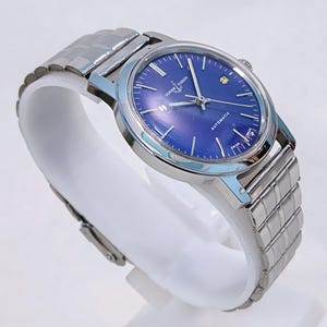 May include: A silver-toned wristwatch with a blue face and a metal band. The watch face has silver hands and markers, with the text "ULYSSE NARDIN AUTOMATIC SWISS MADE". The watch is displayed on a white stand.