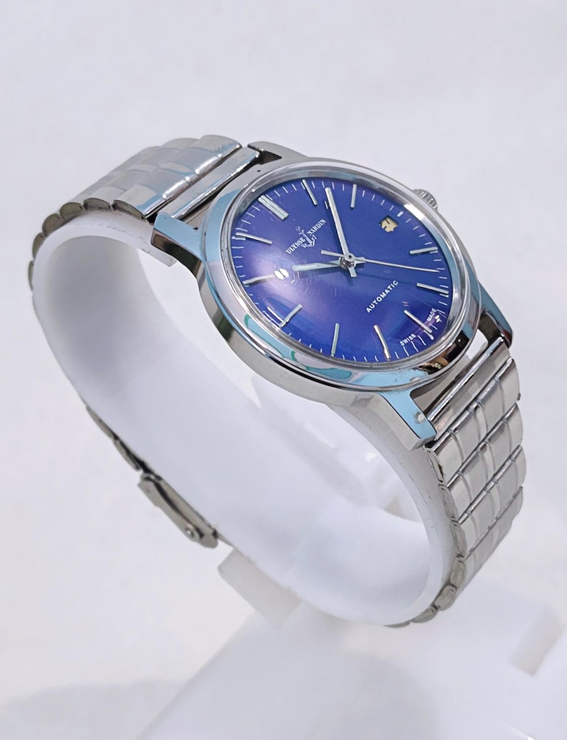May include: A silver-toned wristwatch with a blue face and a metal band. The watch face has silver hands and markers, with the text "ULYSSE NARDIN AUTOMATIC SWISS MADE". The watch is displayed on a white stand.