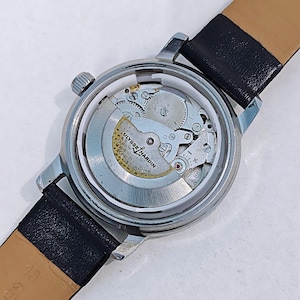 May include: A silver-toned Ulysse Nardin wristwatch with a black leather band. The watch's back is open, revealing the intricate mechanical movement. The watch face is not visible. The band is black on the outside and tan on the inside.