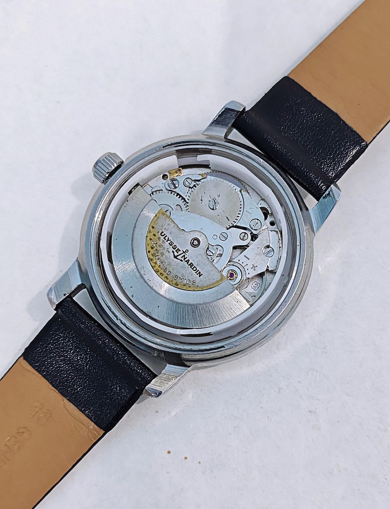 May include: A silver-toned Ulysse Nardin wristwatch with a black leather band. The watch's back is open, revealing the intricate mechanical movement. The watch face is not visible. The band is black on the outside and tan on the inside.