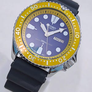 Peut inclure: Montre-bracelet automatique Seiko avec bracelet en caoutchouc noir. La montre a une lunette jaune avec des marquages blancs et un cadran bleu avec des aiguilles et des marqueurs blancs. Le jour est jeudi 22. Résistant à l'eau jusqu'à 150m.