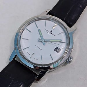 K&ouml;nnte beinhalten: Eine silberfarbene Ulysse Nardin Automatik-Armbanduhr mit wei&szlig;em Zifferblatt und schwarzem Lederarmband. Das Zifferblatt zeigt das Datum und die Worte "AUTOMATIC", "SWISS" und "MADE". Die Zeiger der Uhr sind silberfarben mit leuchtend gr&uuml;ner F&uuml;llung.