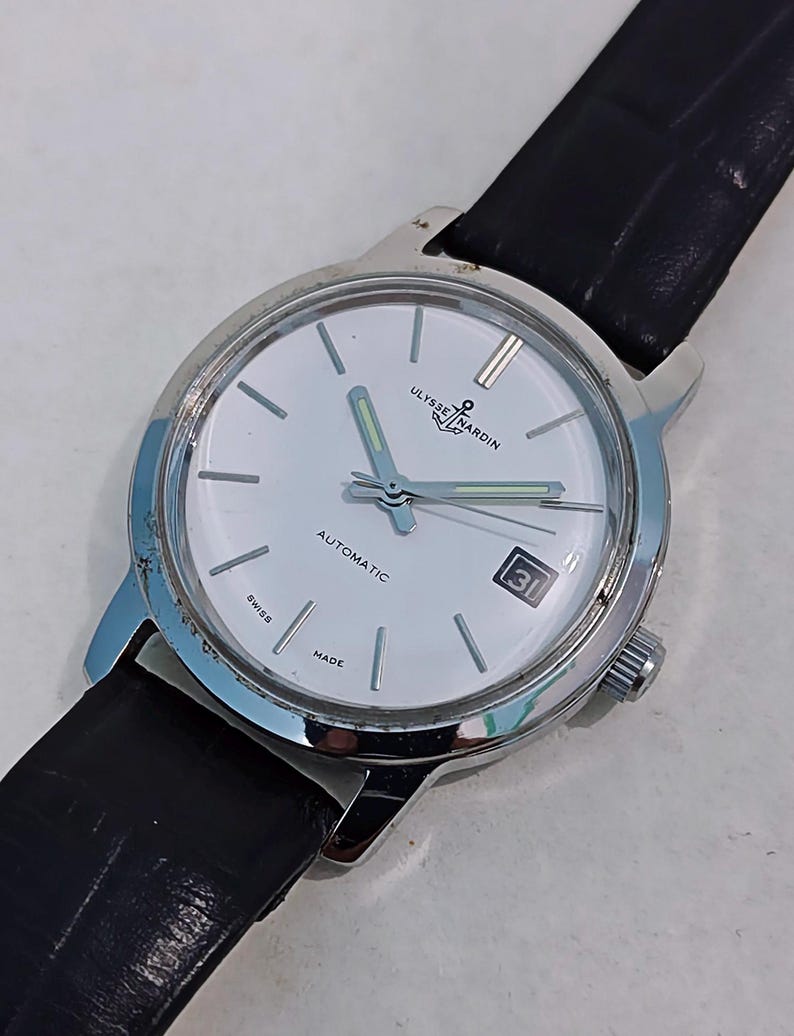 K&ouml;nnte beinhalten: Eine silberfarbene Ulysse Nardin Automatik-Armbanduhr mit wei&szlig;em Zifferblatt und schwarzem Lederarmband. Das Zifferblatt zeigt das Datum und die Worte "AUTOMATIC", "SWISS" und "MADE". Die Zeiger der Uhr sind silberfarben mit leuchtend gr&uuml;ner F&uuml;llung.