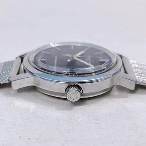 May include: A silver-toned wristwatch with a dark face and a mesh band. The watch has a round face with silver hands and markers. The band is a fine, woven mesh. The watch's side features a textured crown.