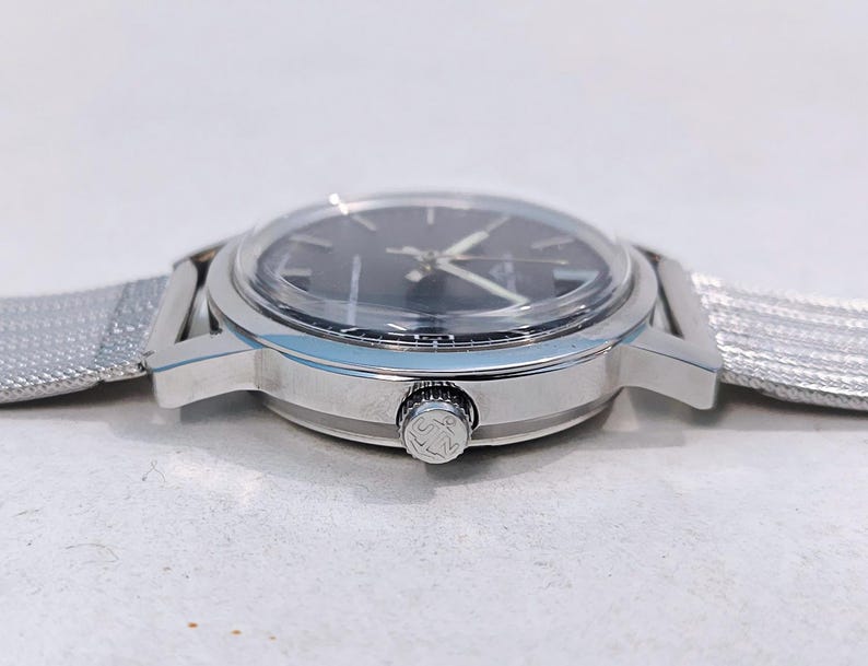 May include: A silver-toned wristwatch with a dark face and a mesh band. The watch has a round face with silver hands and markers. The band is a fine, woven mesh. The watch's side features a textured crown.