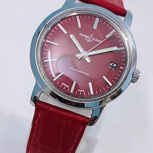 May include: A vintage Ulysse Nardin automatic wristwatch with a red dial and a red leather strap. The watch has a silver-colored case and features the text "AUTOMATIC" and "SWISS MADE". The date is displayed at the 3 o'clock position.