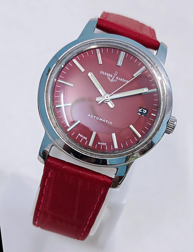 May include: A vintage Ulysse Nardin automatic wristwatch with a red dial and a red leather strap. The watch has a silver-colored case and features the text "AUTOMATIC" and "SWISS MADE". The date is displayed at the 3 o'clock position.