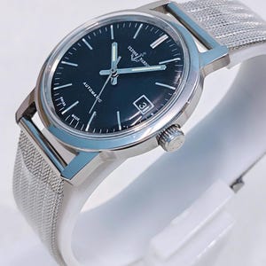 May include: A silver-toned Ulysse Nardin automatic watch with a black face and a mesh band. The watch has a date window at the 3 o'clock position and the words "AUTOMATIC" and "SWISS MADE" are visible on the face.