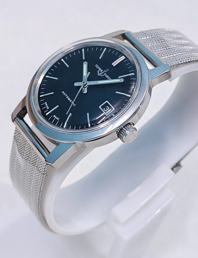 May include: A silver-toned Ulysse Nardin automatic watch with a black face and a mesh band. The watch has a date window at the 3 o'clock position and the words "AUTOMATIC" and "SWISS MADE" are visible on the face.