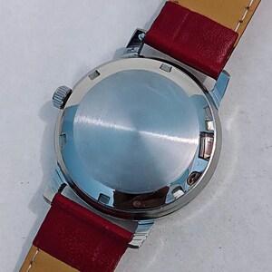 May include: Close-up of a vintage wristwatch. The watch has a silver-colored case and a red leather band with a tan underside. The watch face is not visible. The watch is on a white surface.