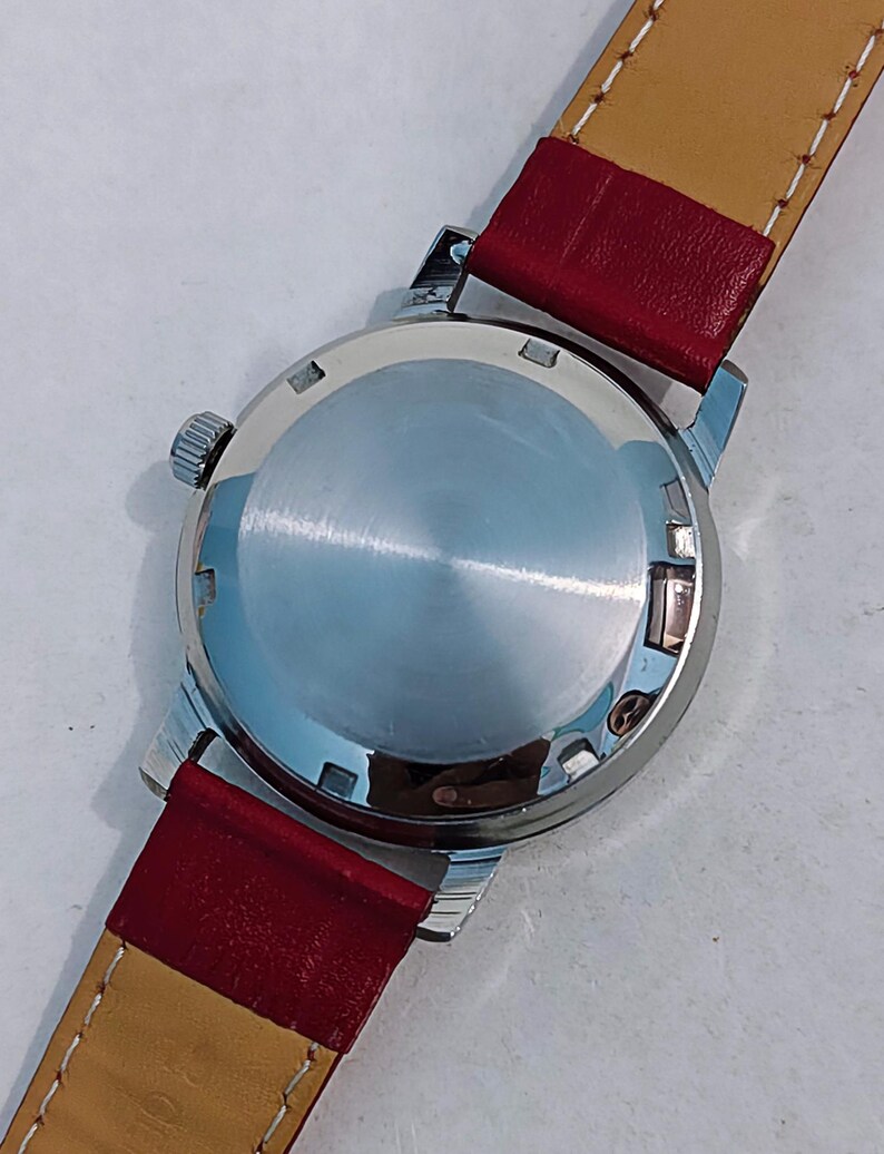 May include: Close-up of a vintage wristwatch. The watch has a silver-colored case and a red leather band with a tan underside. The watch face is not visible. The watch is on a white surface.