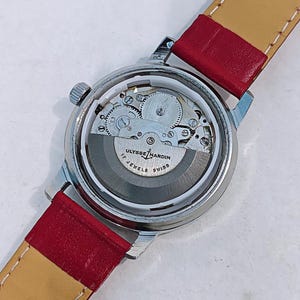 May include: A silver-toned Ulysse Nardin wristwatch with a transparent back, showcasing the intricate inner workings. The watch features a red leather strap with a tan underside. The watch face displays the text "Ulysse Nardin Swiss" and "17 Jewels".