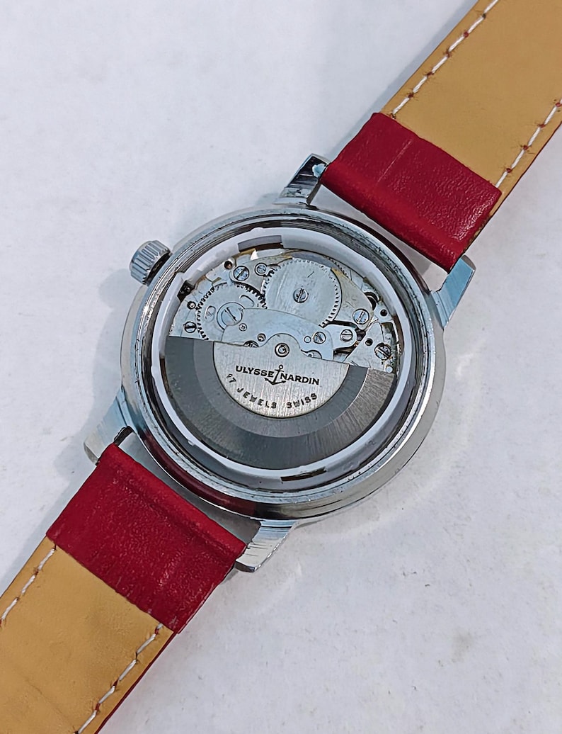 May include: A silver-toned Ulysse Nardin wristwatch with a transparent back, showcasing the intricate inner workings. The watch features a red leather strap with a tan underside. The watch face displays the text "Ulysse Nardin Swiss" and "17 Jewels".
