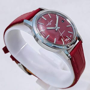 May include: A stainless steel wristwatch with a burgundy dial and a red leather band. The watch face has silver hands and markers, with the text "ULYSSE NARDIN AUTOMATIC SWISS MADE". The watch is displayed on a white stand.