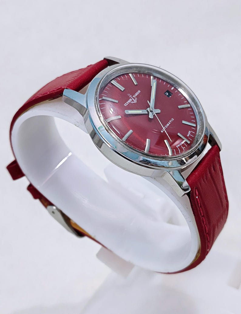 May include: A stainless steel wristwatch with a burgundy dial and a red leather band. The watch face has silver hands and markers, with the text "ULYSSE NARDIN AUTOMATIC SWISS MADE". The watch is displayed on a white stand.