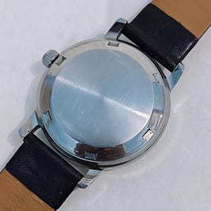 May include: Close-up of a vintage wristwatch, showcasing the watch's stainless steel back and crown. The watch features a black leather strap. The watch's case is silver, and the strap is black with a tan underside.