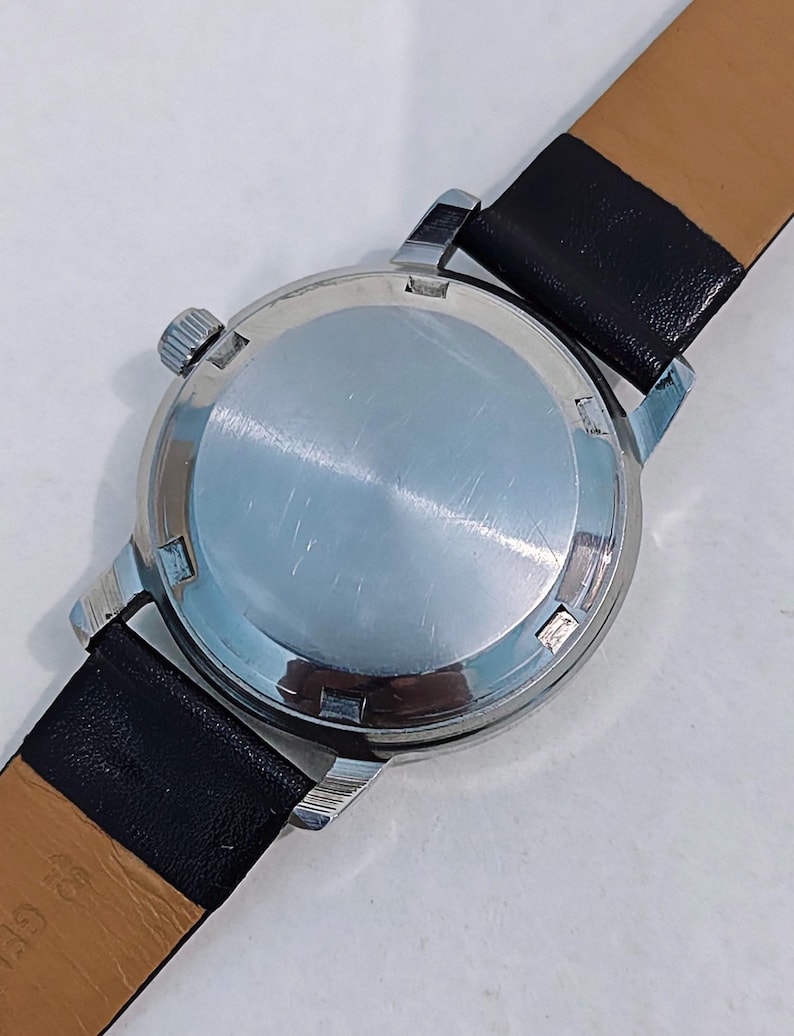 May include: Close-up of a vintage wristwatch, showcasing the watch's stainless steel back and crown. The watch features a black leather strap. The watch's case is silver, and the strap is black with a tan underside.
