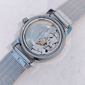 May include: A silver-toned wristwatch with a mesh band, showcasing the intricate internal mechanisms through a transparent back. The watch features the brand name "Ulysse Nardin" and a small crown. The band is made of woven metal.