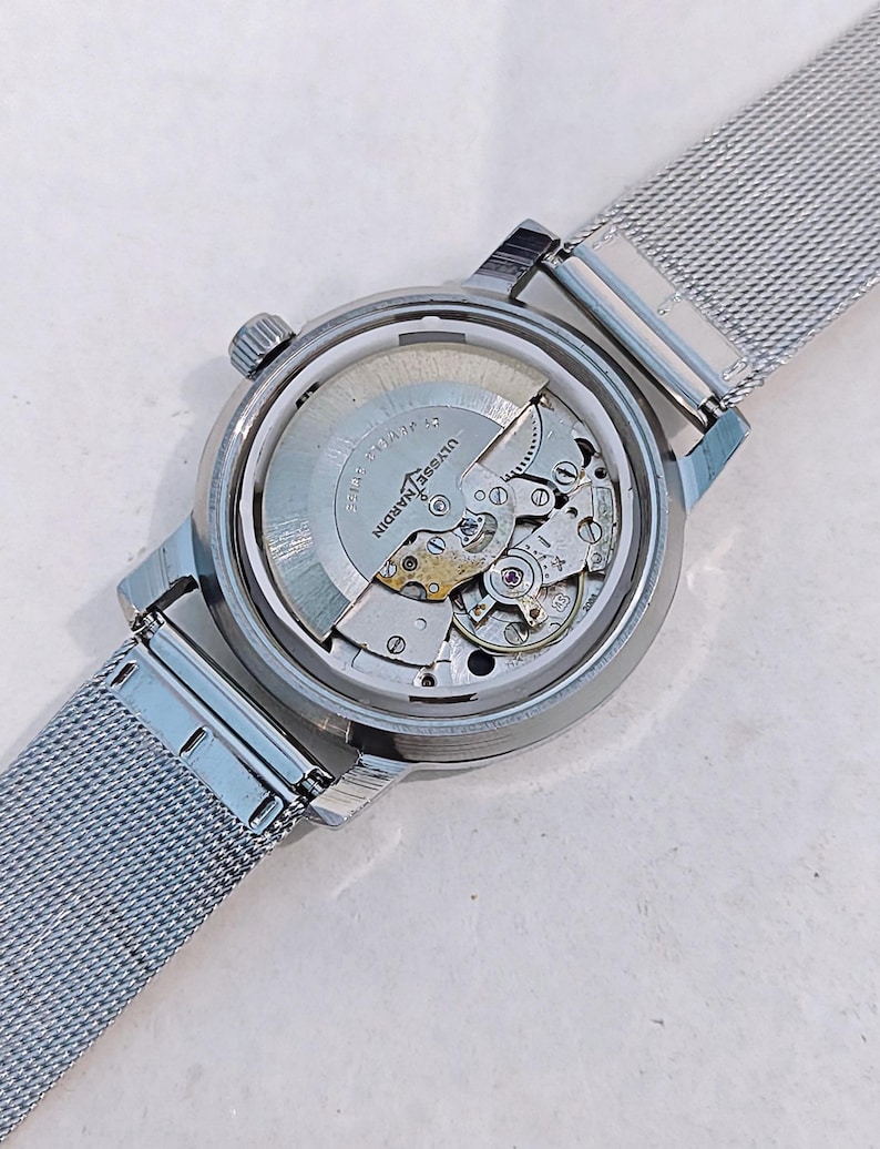May include: A silver-toned wristwatch with a mesh band, showcasing the intricate internal mechanisms through a transparent back. The watch features the brand name "Ulysse Nardin" and a small crown. The band is made of woven metal.