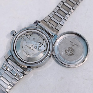 May include: A silver-toned Ulysse Nardin wristwatch with the back cover open, revealing the intricate mechanical movement. The watch has a silver band and a round face. The open back cover displays the words "SWISS ACID INOX" and a logo.