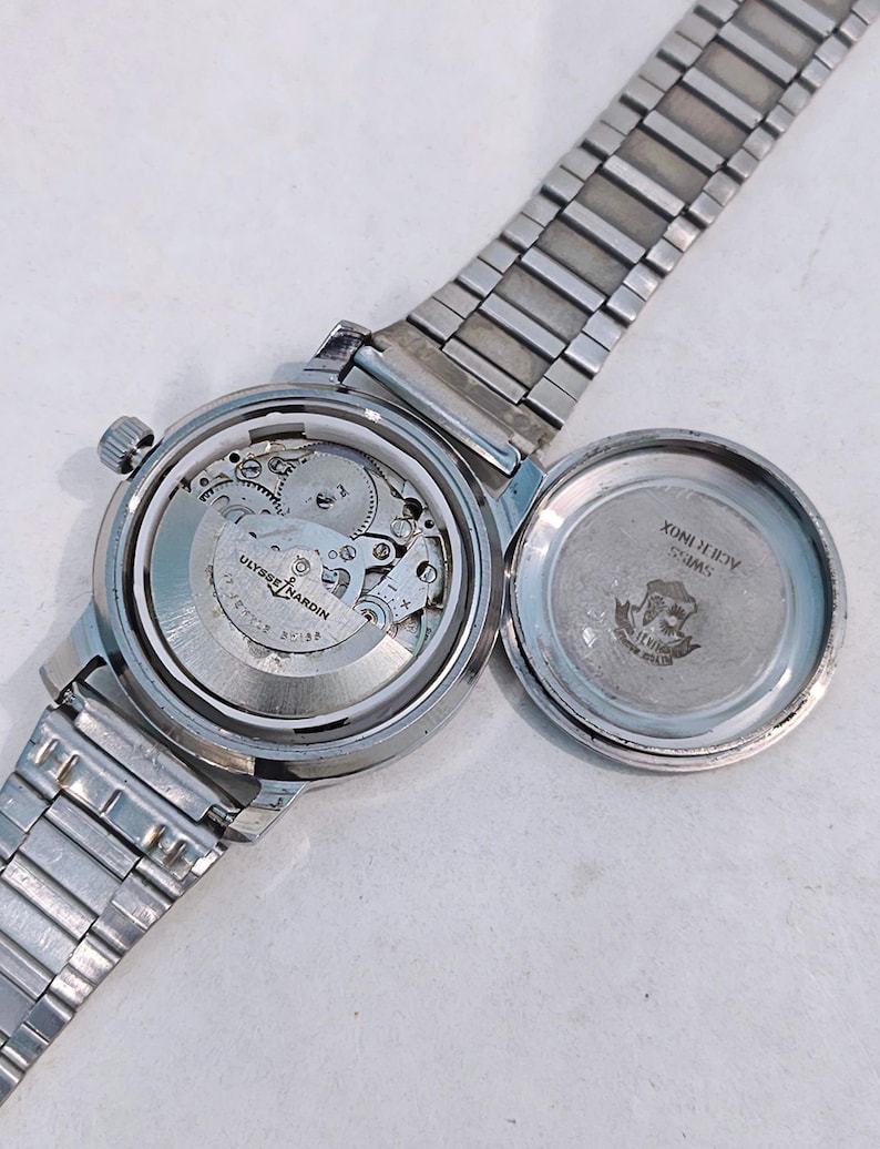 May include: A silver-toned Ulysse Nardin wristwatch with the back cover open, revealing the intricate mechanical movement. The watch has a silver band and a round face. The open back cover displays the words "SWISS ACID INOX" and a logo.