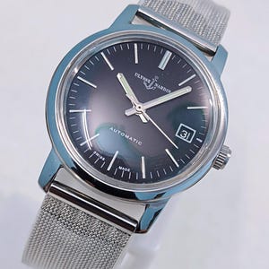 May include: A silver-toned Ulysse Nardin automatic wristwatch with a dark gray face and a mesh band. The watch face displays the date and the words "Automatic", "Swiss", and "Made". The watch has silver hands and hour markers.