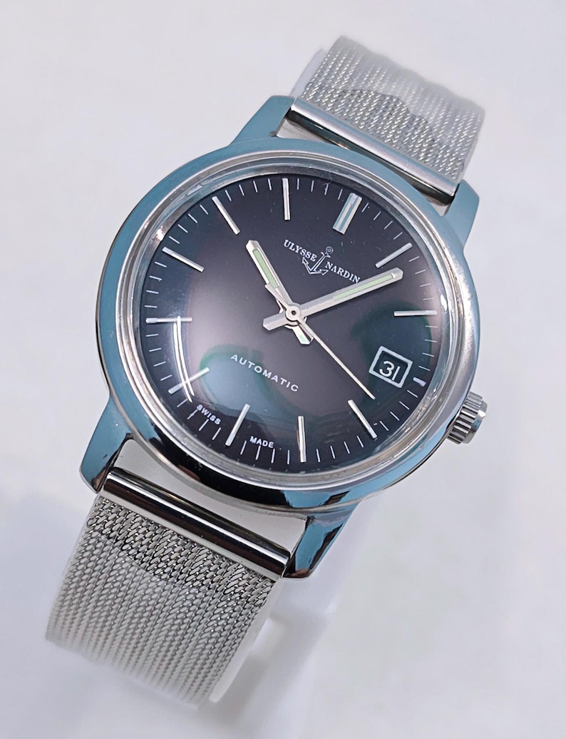 May include: A silver-toned Ulysse Nardin automatic wristwatch with a dark gray face and a mesh band. The watch face displays the date and the words "Automatic", "Swiss", and "Made". The watch has silver hands and hour markers.