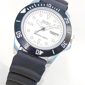 May include: A Seiko automatic diver's watch with a white face, black bezel, and black rubber strap. The watch face displays the day and date, with luminous hands and markers. The bezel has markings for diving, and the watch is water-resistant to 200 meters.