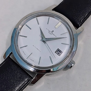 May include: A silver-toned Ulysse Nardin automatic wristwatch with a white face, black leather band, and date display. The watch features silver hands and markers, and the words "SWISS MADE" are visible.