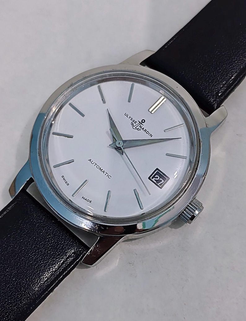 May include: A silver-toned Ulysse Nardin automatic wristwatch with a white face, black leather band, and date display. The watch features silver hands and markers, and the words "SWISS MADE" are visible.
