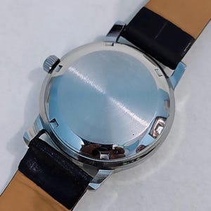 K&ouml;nnte beinhalten: Eine silberfarbene Armbanduhr mit schwarzem Lederarmband. Die Uhr hat ein rundes Zifferblatt und eine kleine Krone. Der polierte R&uuml;cken der Uhr ist reflektierend. Das Armband ist au&szlig;en schwarz und innen beige.