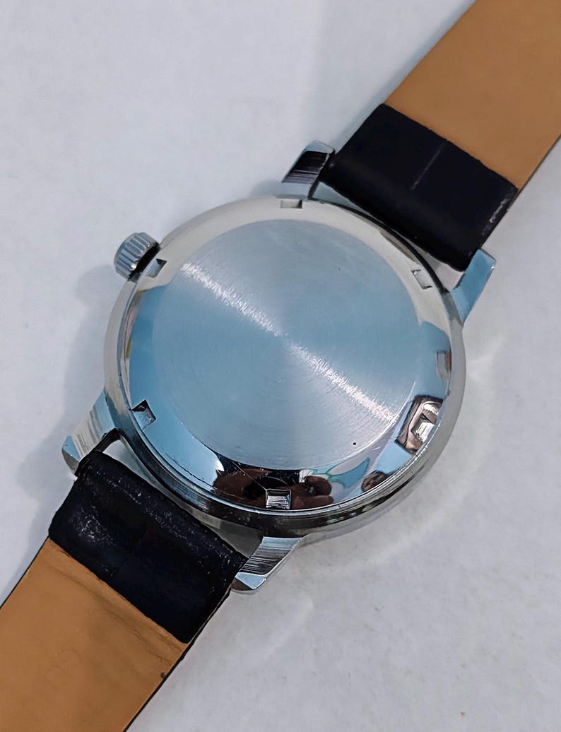 K&ouml;nnte beinhalten: Eine silberfarbene Armbanduhr mit schwarzem Lederarmband. Die Uhr hat ein rundes Zifferblatt und eine kleine Krone. Der polierte R&uuml;cken der Uhr ist reflektierend. Das Armband ist au&szlig;en schwarz und innen beige.